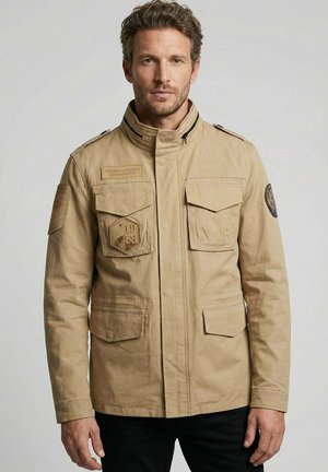 Homme portant une veste style militaire beige avec plusieurs poches avant et des écussons brodés, debout devant un fond uni.