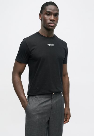 Μαύρο βαμβακερό μπλουζάκι με κοντά μανίκια που φέρει το λογότυπο "VERSACE" σε λευκό, συνδυασμένο με γκρι υφασμάτινο παντελόνι, στέκεται μπροστά σε ουδέτερο φόντο.