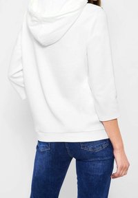 Sudadera blanca con capucha y textura suave, de mangas tres cuartos y con ribete en el dobladillo, combinada con jeans ajustados de color azul oscuro.