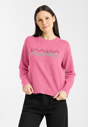 Woman with straight dark hair wearing a pink sweater with the word "ICONIC" and black pants, standing against a white background.