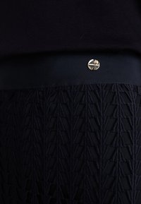 Donker marineblauwe stof met een gestructureerd geometrisch patroon. Een gladde tailleband heeft een klein metallic logo detail.