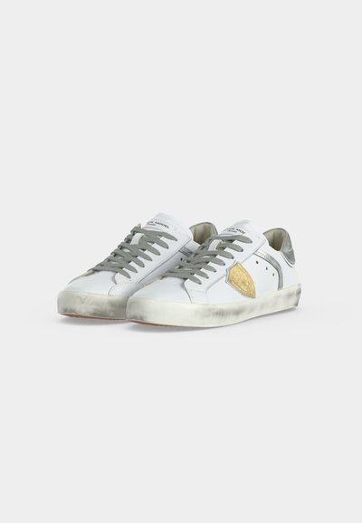 Λευκά sneakers χαμηλού κόμβου με γκρι κορδόνια, ασημένια φτέρνα, κίτρινο πλαϊνό έμβλημα και ελαφρώς βρώμικες σόλες, παρουσιασμένα δίπλα δίπλα σε απλό φόντο.