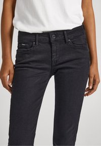 Jeans slim fit negros combinados con una blusa blanca metida por dentro, manos relajadas a los lados, visible botón, detalles de los lazos del cinturón y bolsillos delanteros.