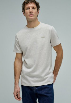 Hombre con cabello rizado que lleva una camiseta de manga corta gris claro y vaqueros azul oscuro, de pie frente a un fondo liso.