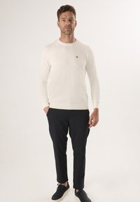 Kremowy sweter z bawełny z prążkowanym okrągłym dekoltem, długimi rękawami i małym brązowym logo na piersiach, zestawiony z ciemnymi spodniami slim fit i czarnymi trampkami.