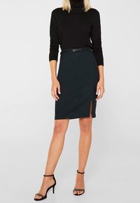 Schwarzer Rollkragenpullover kombiniert mit einem dunkelgrünen Bleistiftrock, der einen seitlichen Schlitz und einen dünnen schwarzen Gürtel hat. Schwarze Riemchen-Heels runden den Look ab.