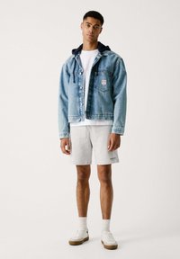 Lichtblauwe spijkerjacket met knoopsluiting aan de voorkant en een patch, gedragen over een wit T-shirt en grijze shorts, aangevuld met witte sneakers.