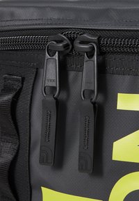 Sac noir avec double fermeture éclair noire portant la marque YKK, en tissu texturé, et détail de logo jaune vif sur la surface.