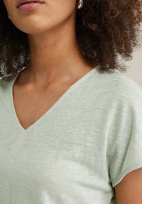 Haut vert clair à col en V avec un tissu doux et texturé et des manches courtes. L'encolure est subtilement inclinée, mettant en valeur un design décontracté.