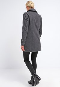 Blonde vrouw die wegloopt in een donkergrijze knielange jas, zwarte smalle broek en zwarte enkellaarsjes tegen een witte achtergrond.