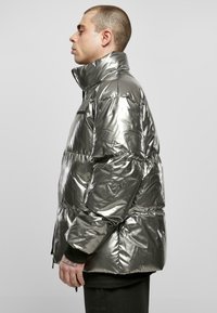 Veste rembourrée argentée métallique avec un col haut, des sections matelassées, des poches latérales et une fermeture éclair. Texture lisse et réfléchissante sur toute la surface.