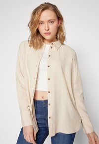 Beige linen shirt with a button-up front, collar, and long sleeves; paired with a white top and blue jeans, showcasing a relaxed fit.