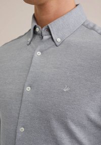 Primer plano de un hombre que lleva una camisa de botones gris claro con cuello abotonado y un sutil logo bordado en el pecho.
