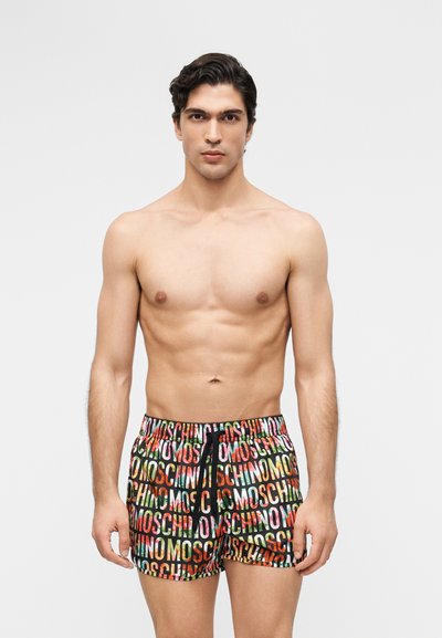 Young man with toned physique wearing colorful shorts patterned with "MOSCHINO" text, standing against plain light background.