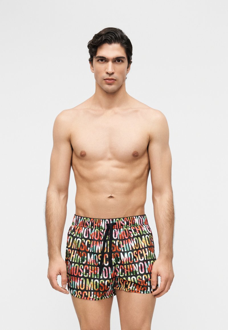 Jeune homme à la silhouette musclée portant un short coloré orné du texte "MOSCHINO", debout devant un fond clair uni.
