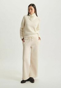 Pull crème côtelé avec un col haut et un pantalon beige à cordon. Tissu doux. Le modèle porte des chaussures sombres. Coupe simple et décontractée.