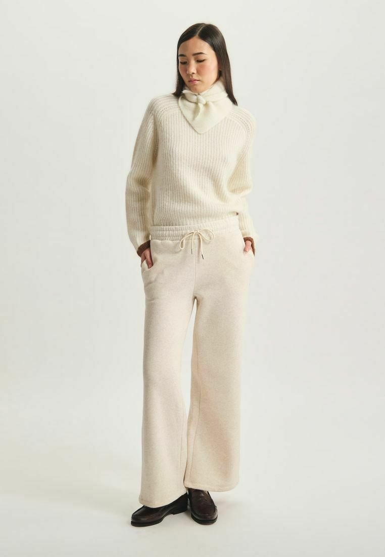 Pull crème côtelé avec un col haut et un pantalon beige à cordon. Tissu doux. Le modèle porte des chaussures sombres. Coupe simple et décontractée.