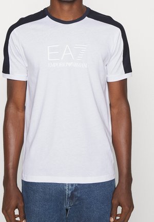 Mann trägt weißes T-Shirt mit schwarzen Schulterstreifen und "EA7 Emporio Armani"-Logo, kombiniert mit blauen Jeans.