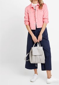 Chaqueta de mezclilla rosa claro con dos bolsillos en el pecho, combinada con pantalones de pierna ancha en color navy. Mochila gris claro con tapa y correas ajustables. Zapatillas blancas.