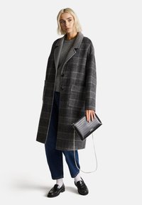 Abrigo de cuadros gris con cuello, botones negros, bolsillos laterales; combinado con un suéter gris, jeans azules y un clutch negro texturizado.