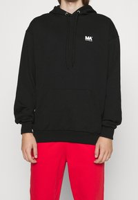 Sort hoodie lavet af blødt stof, med en kængurulomme, snore og et lille hvidt logo på brystet. Båret med røde bukser.