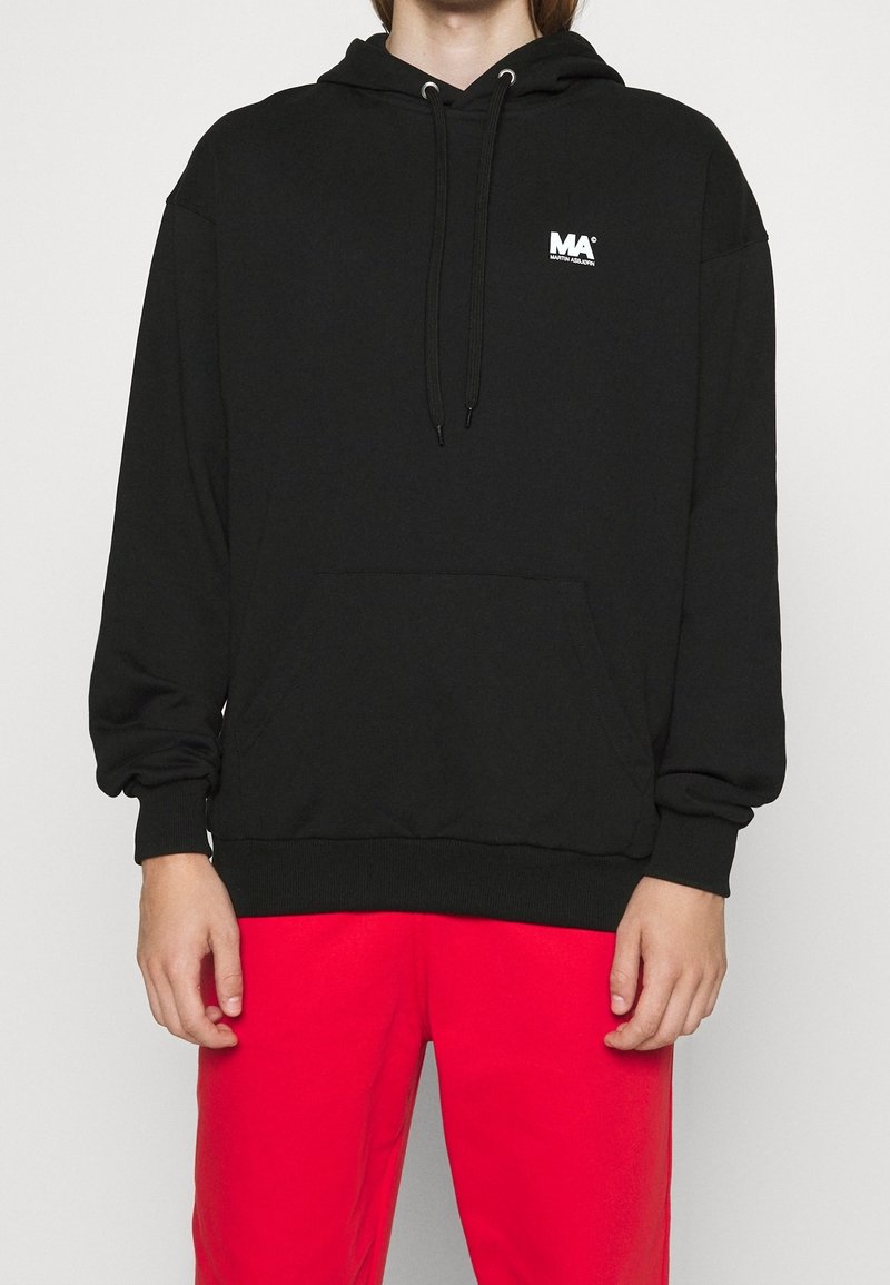 Sort hoodie lavet af blødt stof, med en kængurulomme, snore og et lille hvidt logo på brystet. Båret med røde bukser.