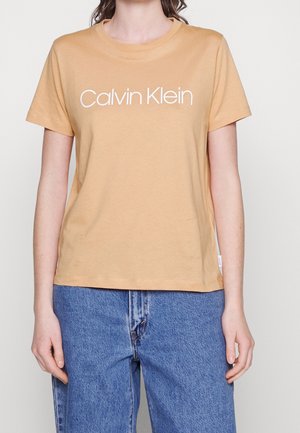 Kobieta ubrana w beżowy t-shirt Calvin Klein z krótkim rękawem i niebieskie dżinsy, stojąca na tle jednolitego jasnego tła.