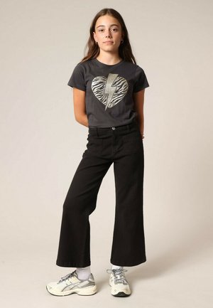 T-shirt gris avec un motif graphique en forme de cœur éclair en or et motif zèbre, associée à un jean noir taille haute à jambes larges et des baskets de couleur claire.