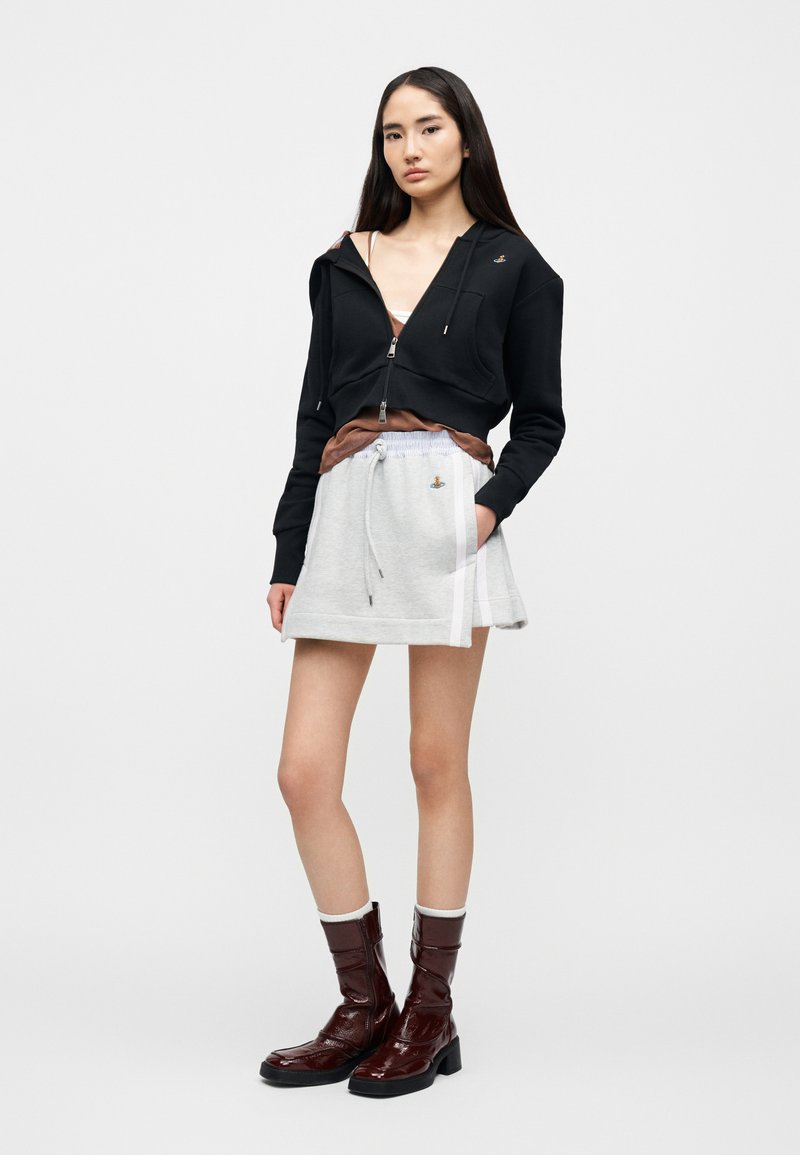 Sweat à capuche noir zippé, jupe grise avec cordon de serrage, et bottines en cuir verni marron à talon carré. Look superposé avec des textures athlétiques.