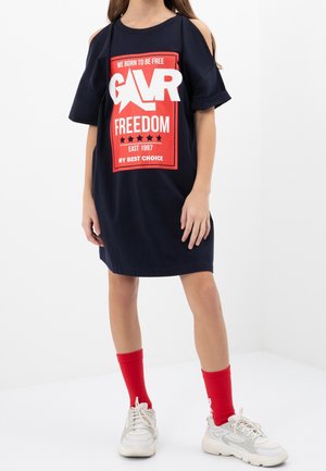 Girl wearing a navy blue cut-out shoulder dress with red and white graphic text, red socks, and white sneakers, standing against white background.