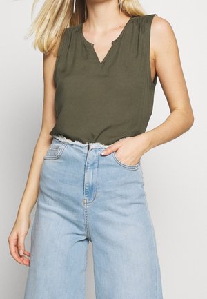 Woman wearing sleeveless olive green top with a V-notch neckline tucked into high-waisted light blue jeans with frayed waistband.