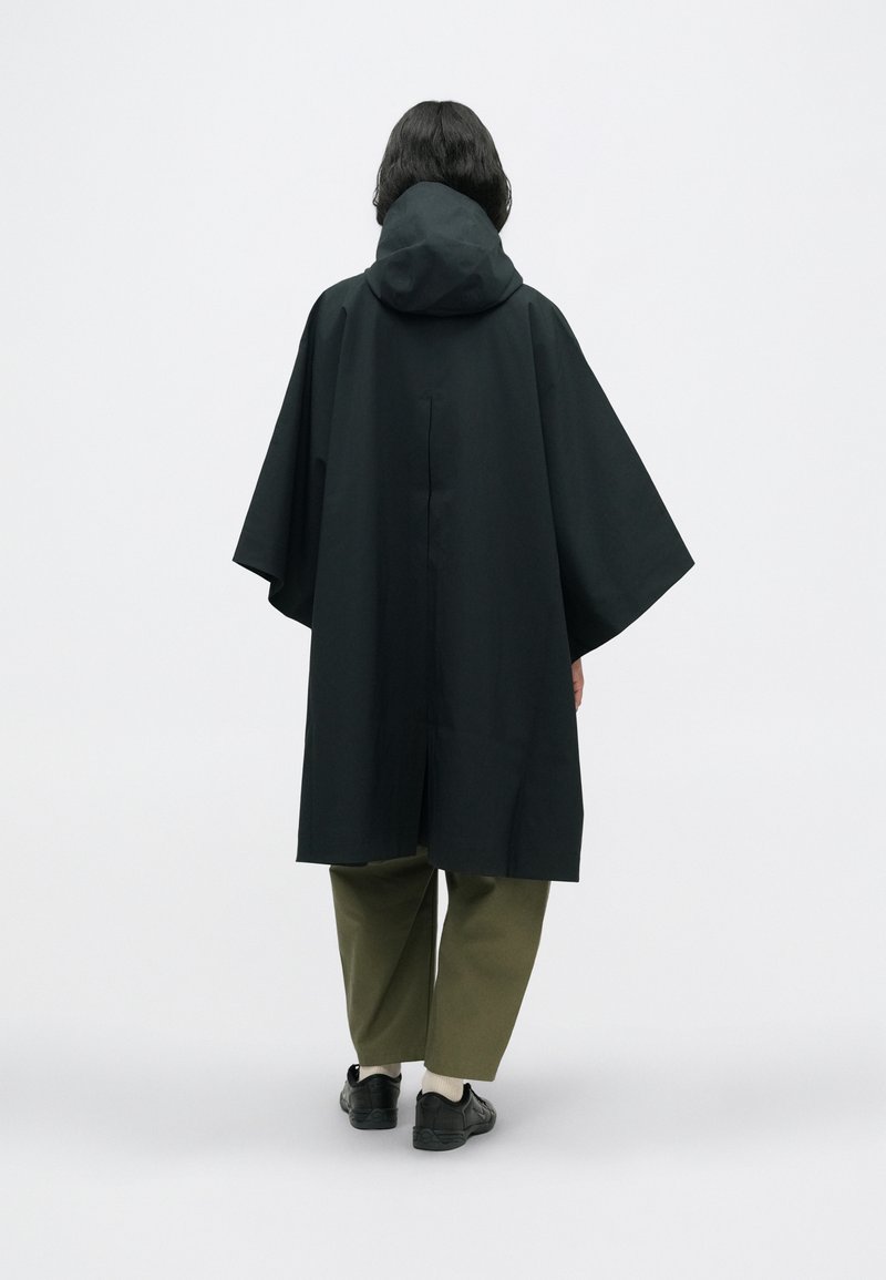 Black hooded poncho with wide sleeves and a straight cut. Features a back ventilation slit. Styled with olive green trousers and black shoes.