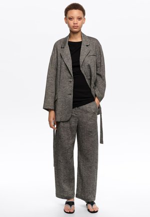 Persona joven de pie vistiendo pantalones anchos grises con textura y una blazer oversize a juego sobre una camiseta negra con sandalias negras de punta abierta.