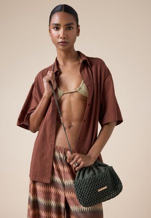 Femme aux cheveux tirés en arrière portant une chemise marron ouverte, un haut de bikini beige, un pantalon à motifs, et tenant un sac à main tressé vert foncé.