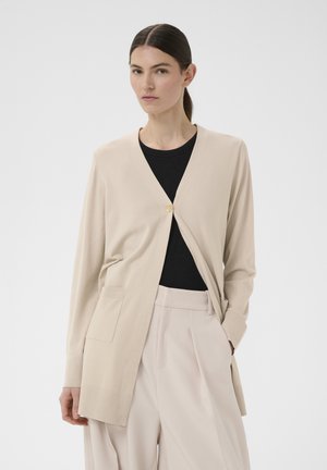 Frau trägt eine beige lange Strickjacke mit einem einzelnen Knopf, ein schwarzes Oberteil und beige weite Hosen, steht vor einem schlichten hellen Hintergrund.