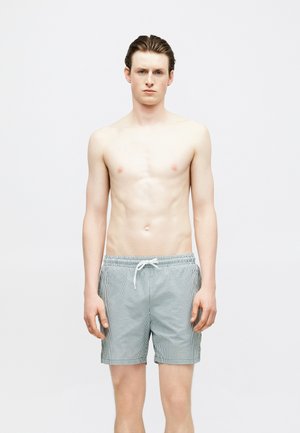 Jeune homme aux cheveux bruns courts portant un short de bain gris et blanc à rayures, debout devant un fond blanc uni.