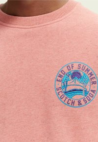 Närbild av en rosa prickig sweatshirt med en blå cirkulär lapp som säger "End of Summer Scotch & Soda" och föreställer ett berg och ett stående djur.