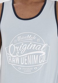 Top fără mâneci de culoare albastru deschis, realizat din bumbac, cu un decolteu nervurat navy și un mare logo grafic alb care spune "Original Raw Denim Co."
