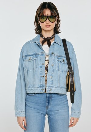 Młoda kobieta nosząca oversize'ową, jasnoniebieską kurtkę jeansową, niebieskie dżinsy, duże zielone okulary przeciwsłoneczne, czarną bandankę na szyi oraz czarną torebkę na ramię.