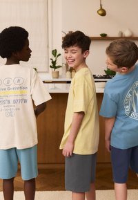 Tre børn står sammen iført t-shirts i gult, beige og lyseblåt, kombineret med shorts i grå og blå. Teksturerne virker bløde.