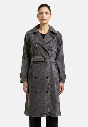 Kvinne som står vendt fremover, iført en mørkegrå, midtlang dobbelspent trenchcoat med belte i midjen og brede slag.