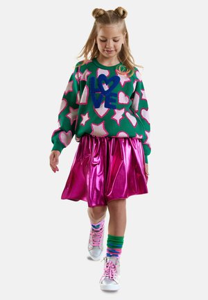 Niña pequeña caminando, vistiendo un suéter verde con estrellas rosas y el texto "LOVE", una falda metálica rosa, calcetines coloridos y zapatillas plateadas.