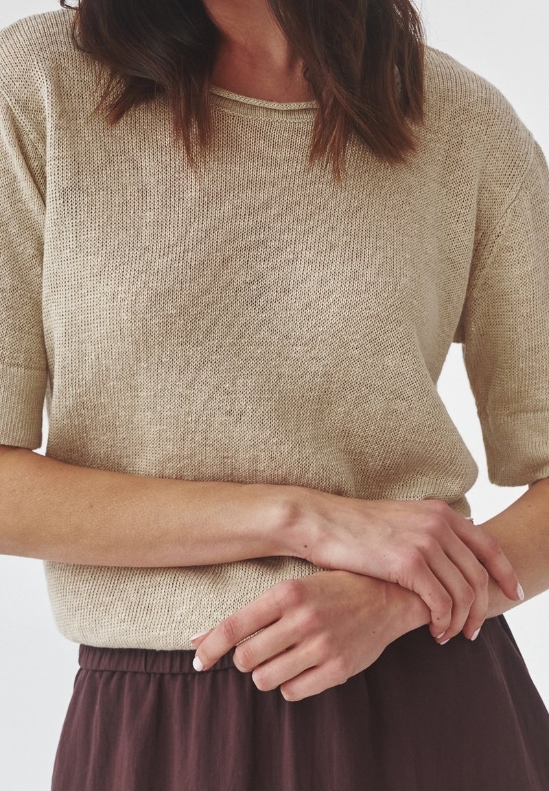 Jersey de punto beige con mangas cortas, escote redondo y corte ancho. La tela tiene una textura suave con un patrón sutil de tejido abierto.