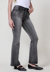 Jeans acampanados grises con un lavado desvanecido, sutiles adornos en los bolsillos y un diseño clásico de cinco bolsillos. Combinados con zapatillas negras.