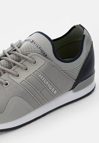 Szara sportowa sneaker z siateczkowego materiału, z szaro-granatowymi akcentami. Zawiera sznurowanie i białą gumową podeszwę. Widoczne logo "Hilfiger".