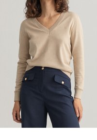 Beige wollen mix trui met een V-hals en ribgebreide manchetten, gecombineerd met marineblauwe broek met gouden knoopaccenten en voorzakken.