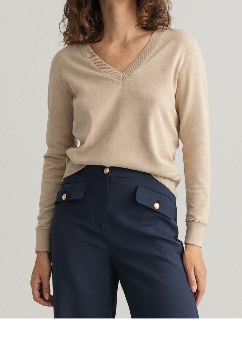 Beige wollen mix trui met een V-hals en ribgebreide manchetten, gecombineerd met marineblauwe broek met gouden knoopaccenten en voorzakken.