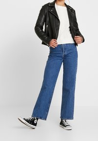 Person wearing a black leather jacket, white cable-knit sweater, blue wide-leg jeans, and black high-top sneakers standing against white background.