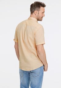 Camisa de manga corta con cuello en tela amarilla clara con un acabado texturizado. Vista trasera que muestra una costura de canesú y mangas remangadas. Se lleva con pantalones vaqueros azules.
