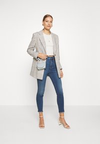 Blazer cinza xadrez com lapelas em pico, top cropped branco, jeans skinny azuis e mala mini cinza claro, complementados por sapatos de salto alto nude.